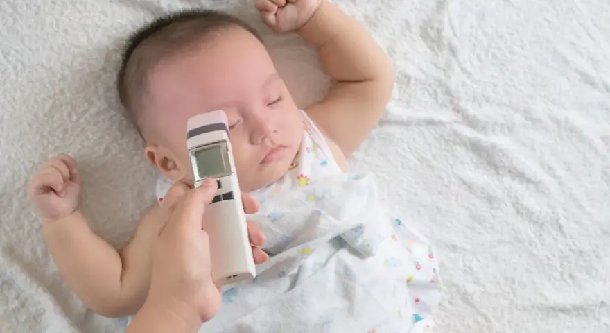 Ręka mierzy niemowlakowi temperaturę na czole termometrem bezdotykowym. Spokojny sen malucha to znak, że prawidłowa temperatura u niemowlaka na czole jest utrzymana.