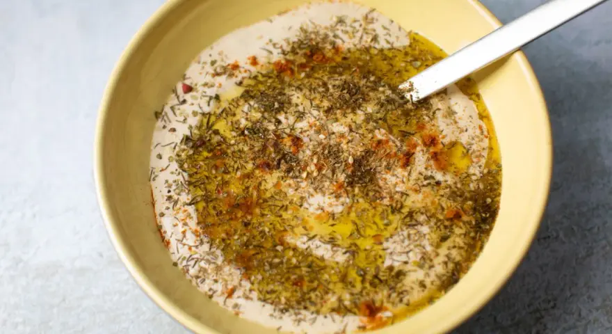 Cremiger Hummus Aufstrich in gelber Schüssel, beträufelt mit Olivenöl und bestreut mit Kräutern und Paprika.