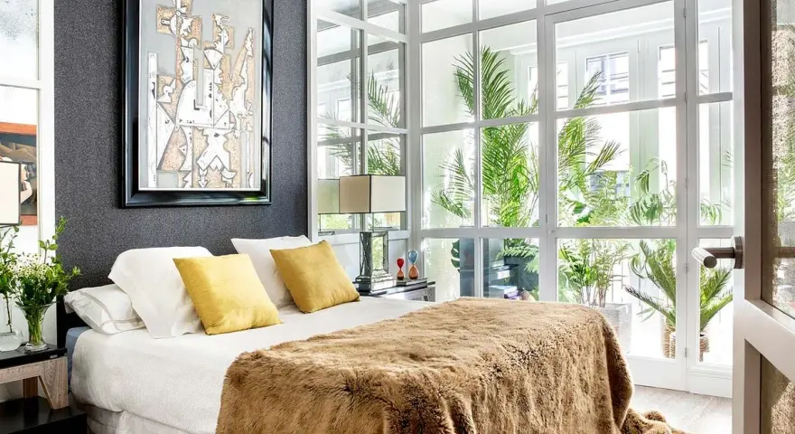 Dormitorios cálidos y modernos con cama mullida, mantas de pelo y gran ventanal con plantas.