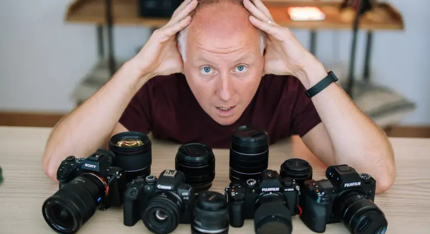 Mężczyzna z rękami na głowie otoczony aparatami różnych marek: Sony, Canon, Fujifilm.