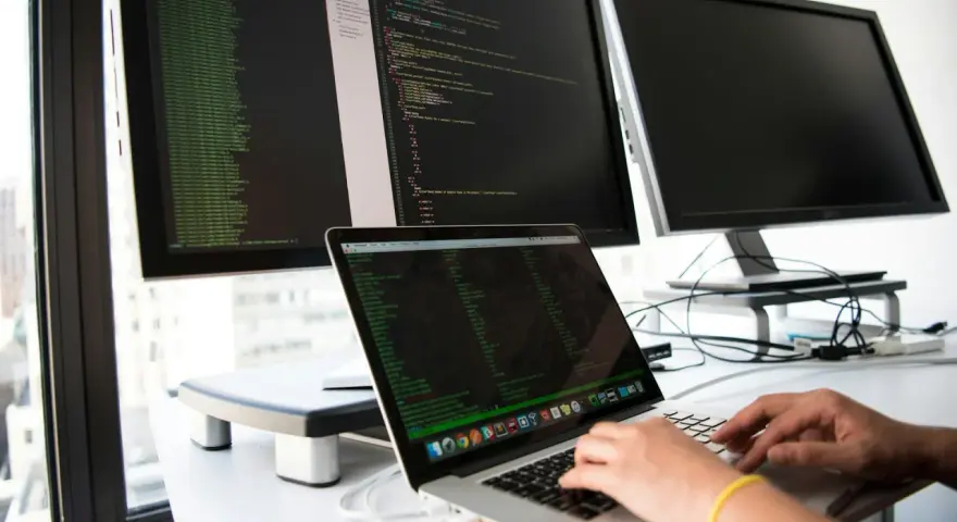 Hands typing on a laptop, surrounded by monitors displaying code. This setup is ideal for developing network infrastructure software.