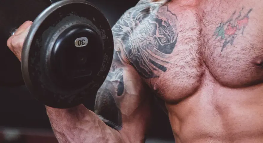Muskulös man med tatueringar lyfter en 30 kg hantel. Frågan är om man kan bygga muskler på kaloriunderskott.