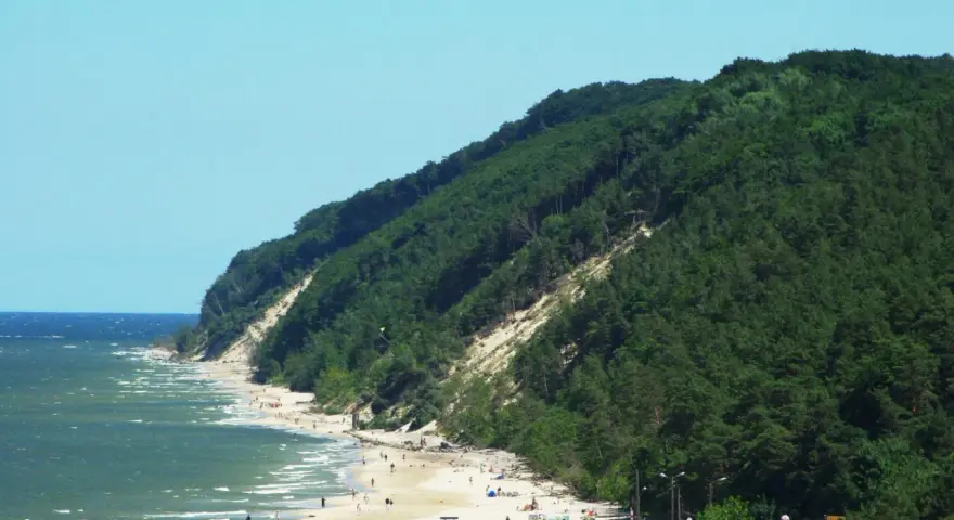 Słoneczna plaża u podnóża zalesionej Kawczej Góry. Woda faluje, a na piasku odpoczywają ludzie.