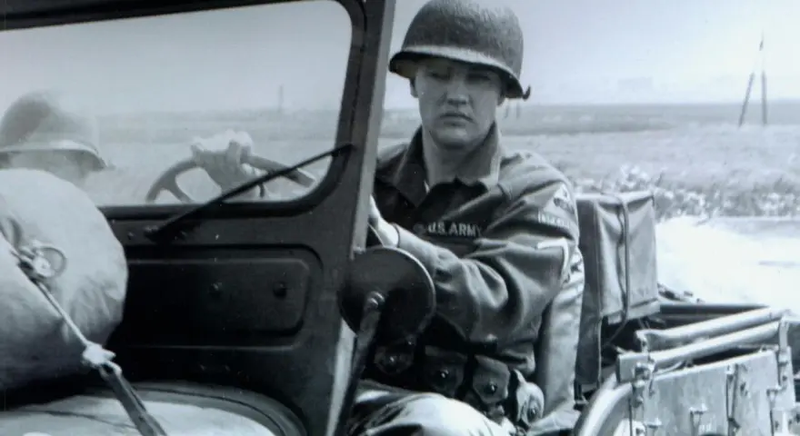 Elvis Presley in seiner US Army Uniform, sitzt am Steuer eines Jeeps.