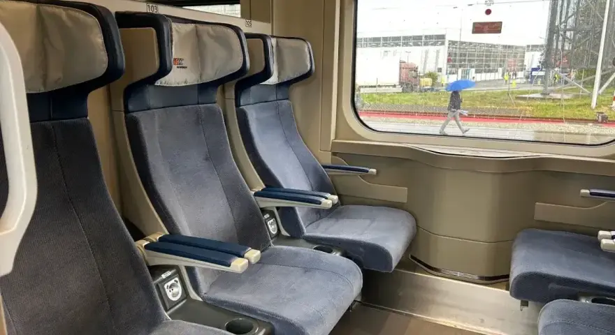 Wygodne siedzenia w wagonie z przedziałami Intercity 2 klasa. Za oknem deszczowa pogoda i przechodzień z niebieskim parasolem.