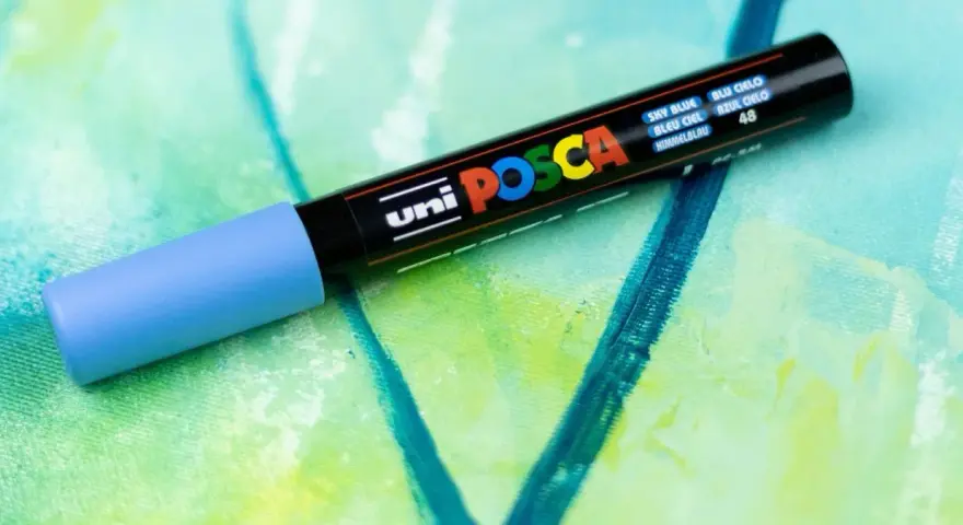 En ljusblå uni POSCA vattenfast tuschpenna ligger på en abstrakt målning i gröna och blå nyanser.