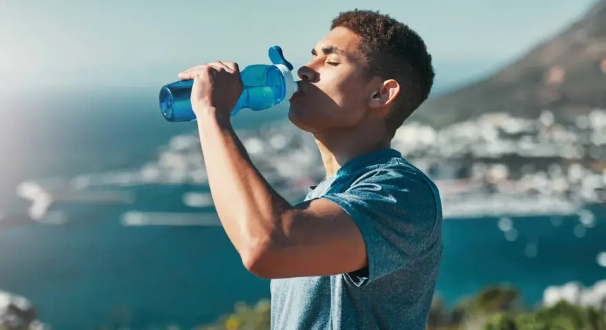 Un jeune homme boit dans une gourde bleue, se réhydratant après l'effort. L'eau, essentielle, contient des électrolytes pour le bon fonctionnement du corps.
