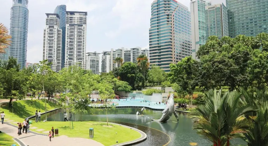 Park z rzeźbą wieloryba i basenem w stolicy Malezji, Kuala Lumpur, otoczony nowoczesnymi wieżowcami.