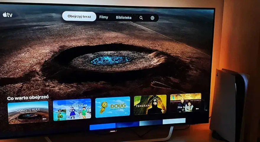 Telewizor z interfejsem Apple TV i konsola PS5 obok. Na ekranie widać menu z filmami i serialami, np. "Ogniste Kule" i "Moja Ewolucja".
