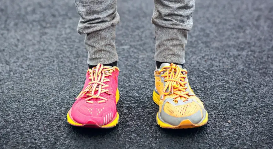 Gambe con pantaloni grigi e scarpe da corsa, una rosa acceso e l'altra arancione, con lacci gialli. La suola scarpa è pronta per correre.