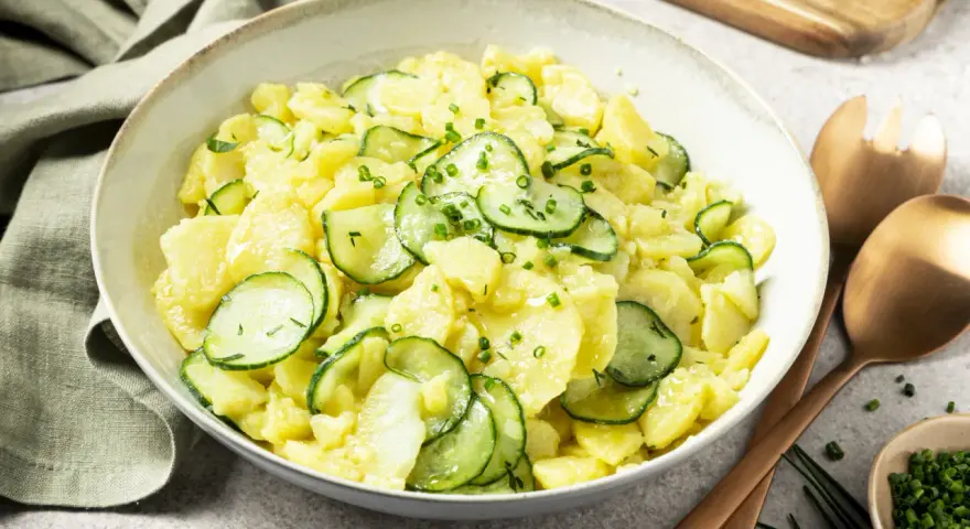 Schüssel mit Kartoffelsalat mit Gurken, garniert mit Schnittlauch. Daneben liegen Servierbesteck und eine kleine Schale mit gehacktem Schnittlauch.