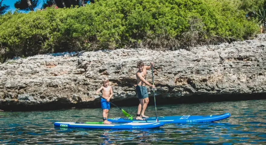 Zwei Jungen auf SUP-Boards, die geradeaus fahren, genießen das klare Wasser vor einer felsigen Küste mit üppiger Vegetation.