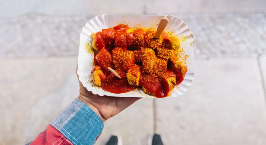 Eine Hand hält eine Portion Currywurst mit original Currywurst Sauce, bestreut mit Currypulver. Ein Holzspieß steckt darin.