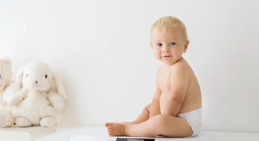 Uśmiechnięty chłopczyk w pieluszce siedzi na wadze. Chyba już czas na większy rozmiar, kiedy pampers jest za mały.