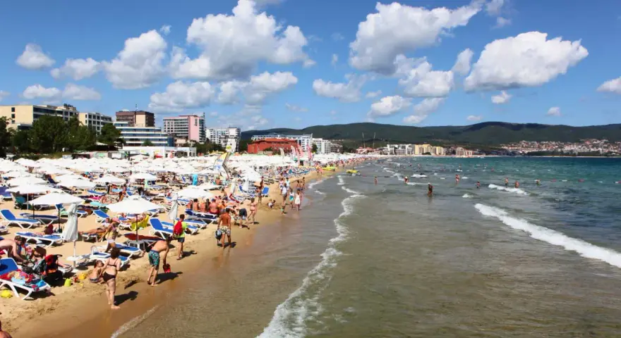 Tłum ludzi wypoczywa na piaszczystej plaży Słoneczny Brzeg, opalając się na leżakach pod białymi parasolami. W tle widać morze i zabudowania kurortu.