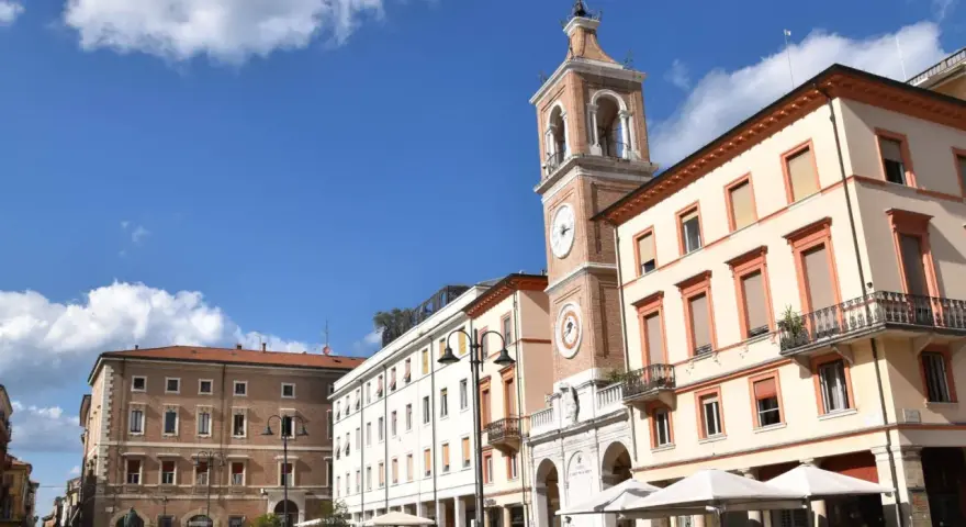 Piazza Tre Martiri a Rimini: un campanile con orologio domina la scena, tra palazzi storici e tavolini all'aperto. Ecco cosa vedere!