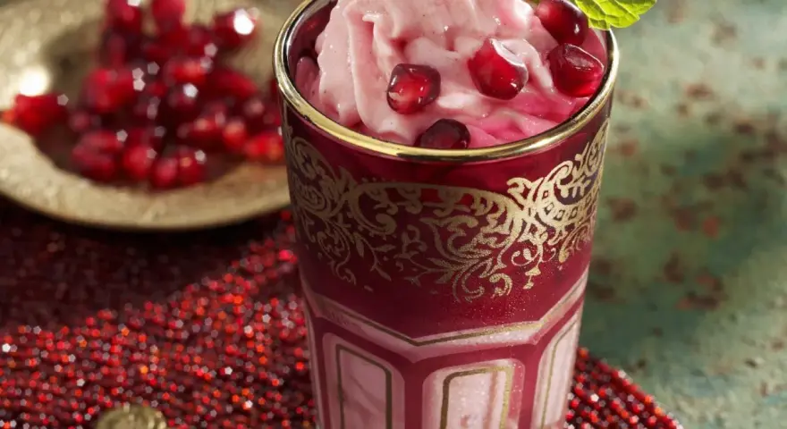 Ein Glas mit cremigem granatapfel eis, garniert mit frischen Granatapfelkernen und Minze. Daneben eine Schale mit Granatapfelkernen.