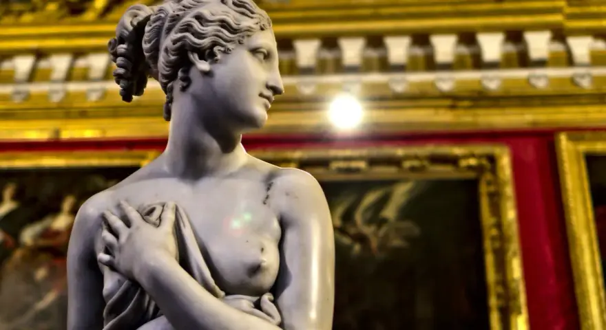 Rzeźba Wenus, dzieło Antonio Corradini, z dłonią na piersi, na tle złoconych ram obrazów.