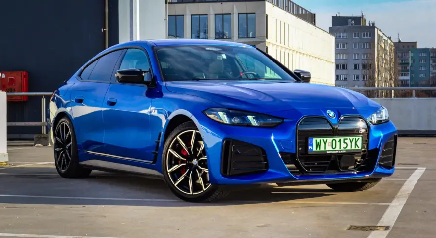 Niebieskie BMW i4 M50 zaparkowane na parkingu, z widokiem na budynki w tle.