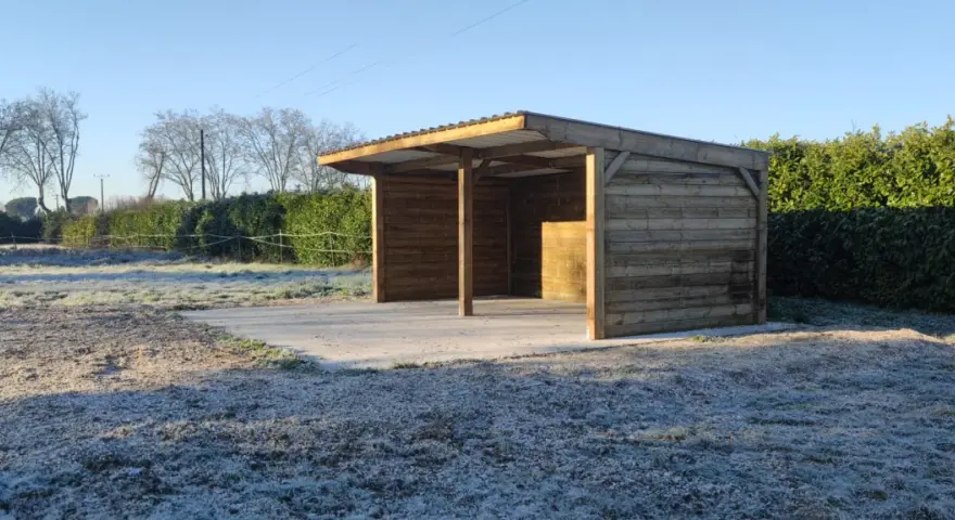 Abri pour chevaux d'occasion en bois, avec toit métallique, sur un sol gelé. Parfait pour protéger vos équidés du froid.