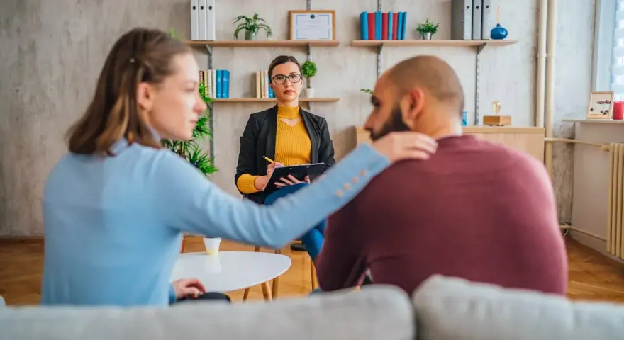 Un couple en consultation avec une psychologue conjugale. La femme pose une main réconfortante sur l'épaule de son partenaire.