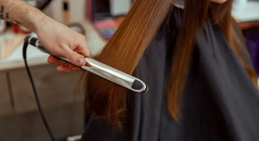 Stylista pokazuje, jak zrobić fale prostownicą, tworząc delikatne loki na długich, brązowych włosach.