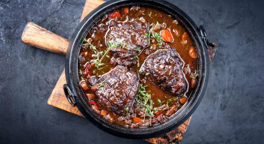 Carrilleras de ternera estofadas en salsa oscura con zanahorias y hierbas, servidas en una cazuela de hierro fundido sobre tabla de madera.