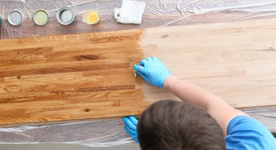 Mani guantate applicano un trattamento per come indurire il legno. Tavolo in legno con diverse lattine di finitura.