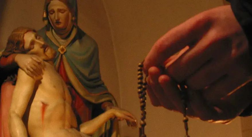 Eine Hand hält einen Rosenkranz, während die Pietà-Statue im Hintergrund zu sehen ist. Ein evangelischer Rosenkranz wird hier in Andacht gehalten.