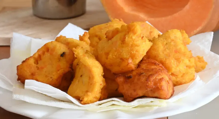 Frittelle di zucca dorate e croccanti, servite su carta assorbente, con una fetta di zucca sullo sfondo.