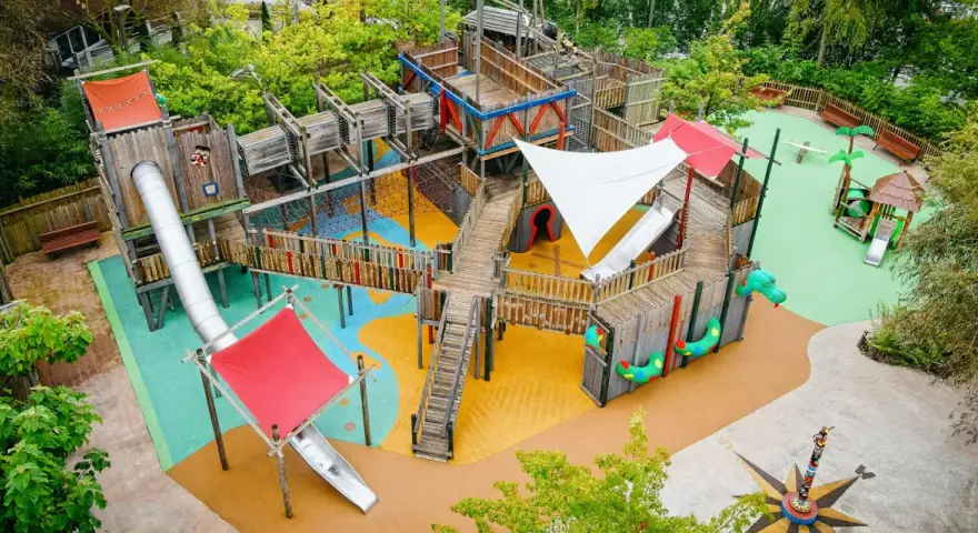 Ein riesiger, mehrstöckiger **Legoland Spielplatz** mit Rutschen, Kletternetzen und Holzbrücken, umgeben von grünen Bäumen.