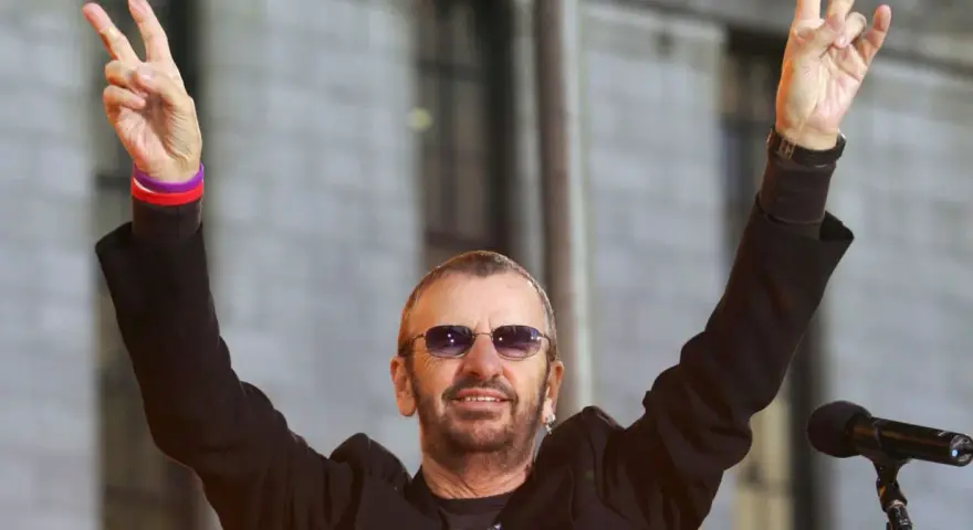 Ringo Starr z The Beatles pozdrawia fanów, unosząc ręce w geście pokoju.