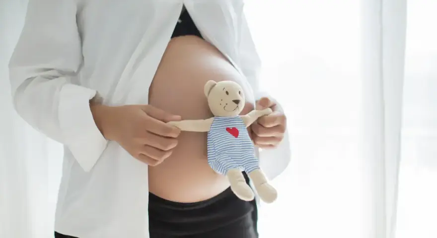 Brzuszek w 21 tygodniu ciąży, otulony białą koszulą. Dłonie trzymają misia w pasiastym ubranku, symbolizującego przyszłe macierzyństwo.