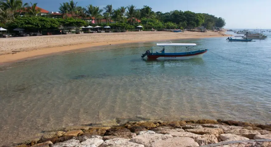 Spokojna zatoka w Sanur na Bali, z łodzią "Water Walk" kołyszącą się na turkusowej wodzie, plażą i palmami w tle.