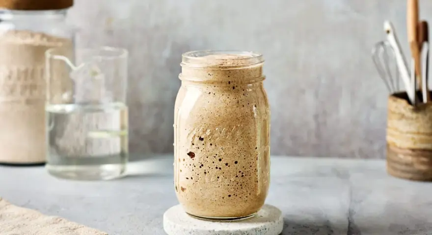 Ein Glas mit sprudelndem Sauerteig, angesetzt mit Dinkelmehl. Daneben ein Glas Wasser und ein Behälter mit Küchenutensilien.