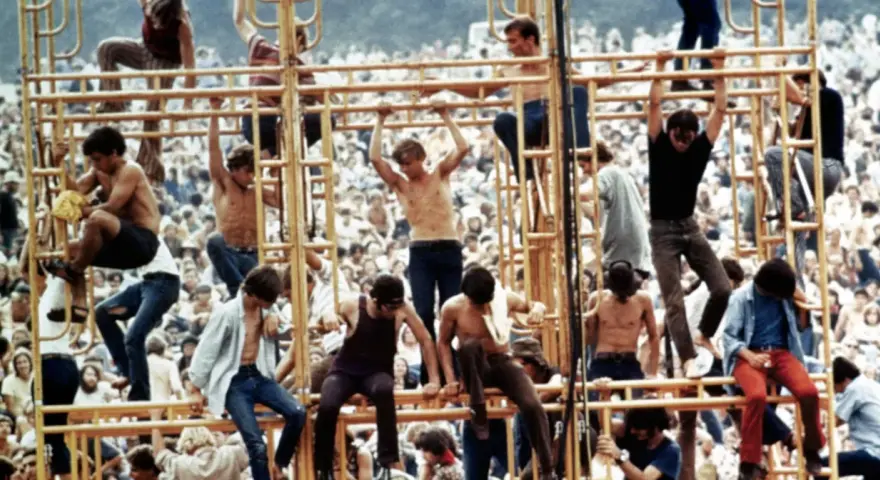 Tłum ludzi na festiwalu, niektórzy wspinają się na konstrukcję. To właśnie woodstock co to za wydarzenie?