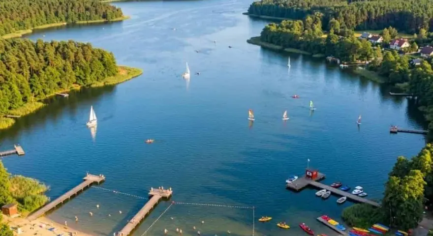 Letnie popołudnie nad jeziorem Kałębie. Żaglówki i kajaki na wodzie, plażowicze na piaszczystym brzegu, otoczonym zielonym lasem.