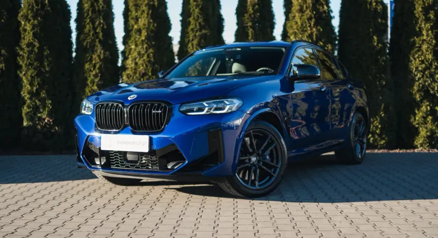 Błyszczący, niebieski suv BMW X4 M Competition zaparkowany przed żywopłotem.
