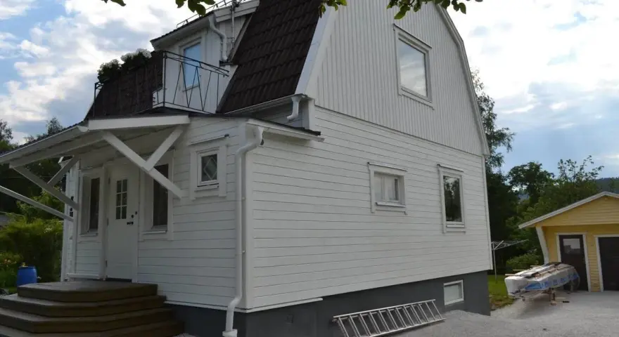 Vitt hus med ny fasad, stegar och material för byta fasad. En gul garagebyggnad syns i bakgrunden.