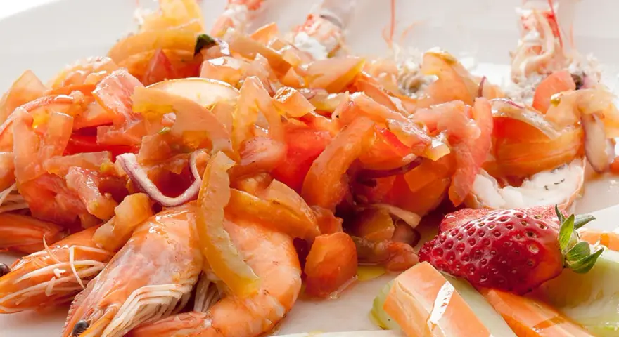 Scampi alla catalana: gamberi succulenti con pomodori freschi, cipolla rossa e una fragola decorativa. Un piatto colorato e invitante.