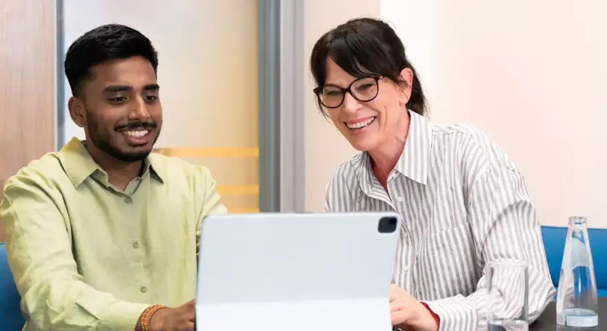 Ein junger Mann und eine Frau lachen über den Bildschirm eines Tablets. Sie sind bei einem Praktikum Studium und scheinen eine gute Zeit zu haben.