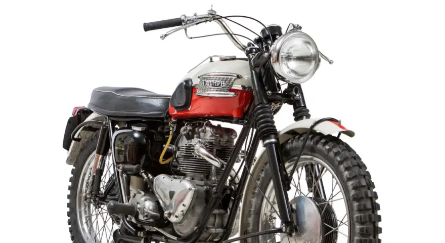 Clásica moto Triumph, modelo scrambler, con tanque bicolor rojo y blanco, motor expuesto y llantas todoterreno.