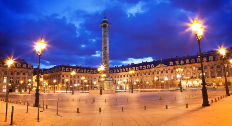 La majestuosa plaza Vendôme se ilumina bajo un cielo nocturno azul intenso, con farolas doradas y la imponente columna en el centro.