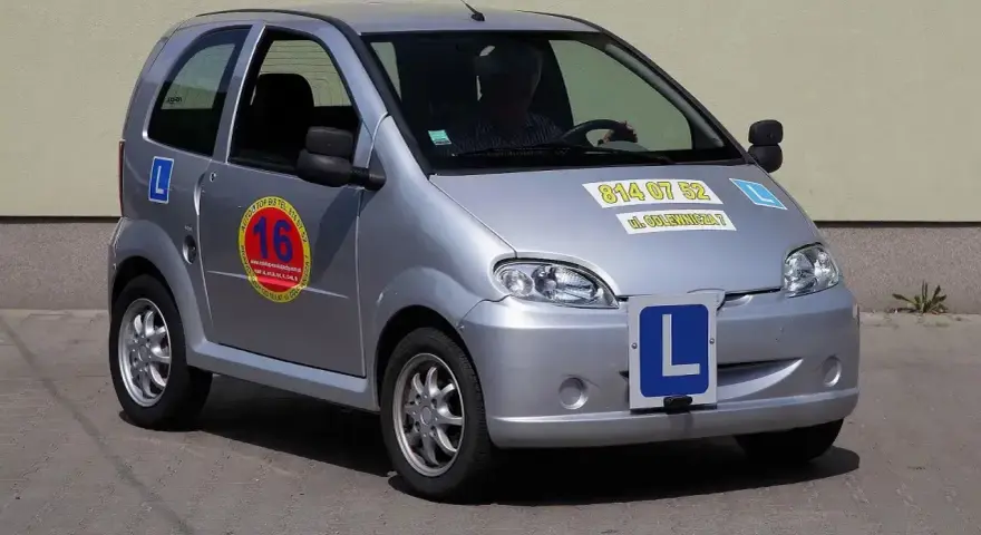 Srebrny samochód do nauki jazdy, kategoria B1. Z przodu i z boku widoczne litery "L", a na drzwiach okrągłe logo szkoły jazdy.