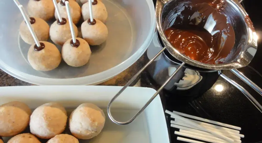 Biskuit-Cake-Pops warten auf ihre Schokoladenhülle. Geschmolzene Schokolade und Stäbchen liegen bereit.