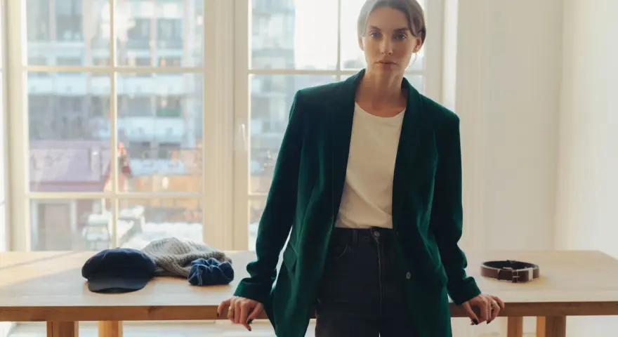 Elegancka kobieta w ciemnozielonej marynarce z aksamitu, stojąca przy drewnianym stole z akcesoriami.
