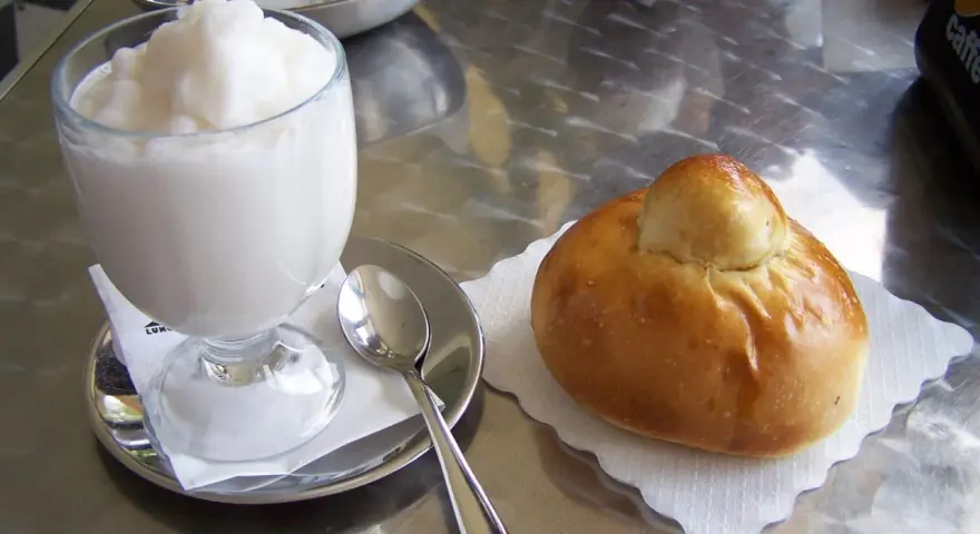 Un bicchiere di granita bianca e un soffice brioche col tuppo su un tavolo metallico.