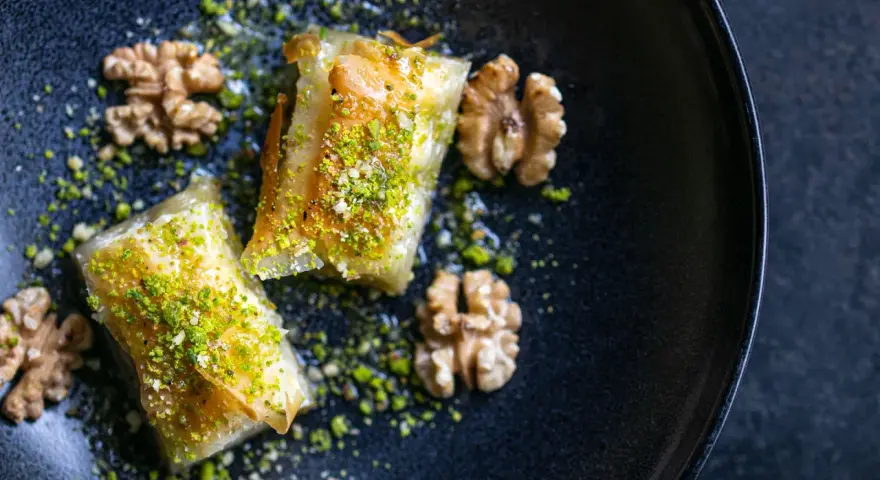 Zwei Stück Baklava, bestreut mit gehackten Pistazien und Nüssen, auf einem dunklen Teller. Ein wahrhaftiges Baklava Rezept Original.
