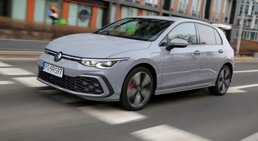 Szary Volkswagen Golf GTE porusza się po miejskiej drodze, z rozmytym tłem budynków i przechodniów.