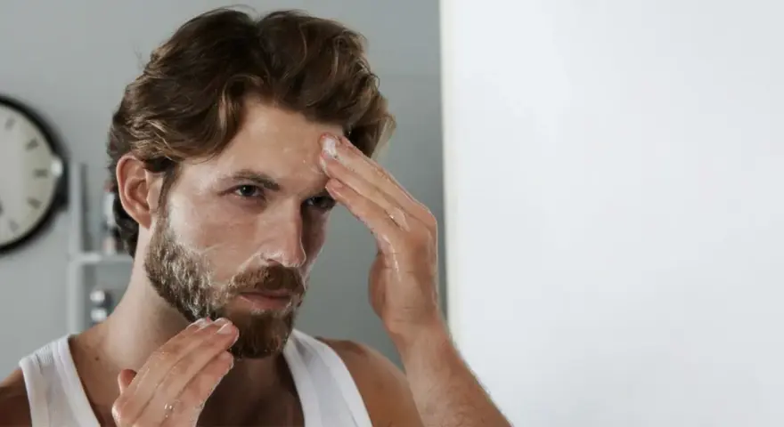 Mann mit unreiner Haut reinigt sein Gesicht mit Schaum. Er blickt in den Spiegel, während er seine Stirn einreibt.